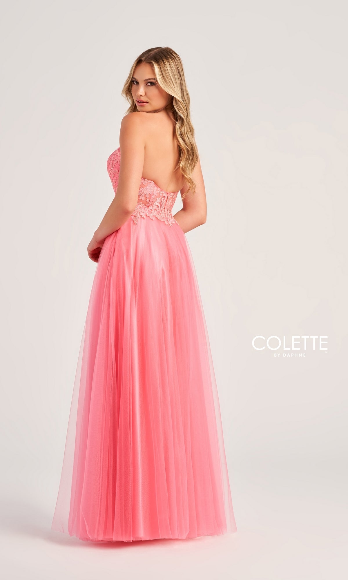 A woman in the Colette Strapless Long A-Line Prom Dress CL5132, featuring a lace bodice and tulle floor-length pink skirt, stands looking over her shoulder. The brand name appears on the right for an elegant finish.